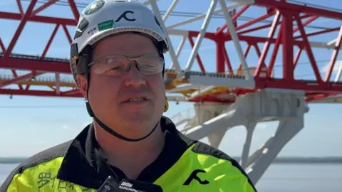 A man standing on a ship wears a white helmet, glasses and a hi-vis jacket. Behind him is a large red and white crane.