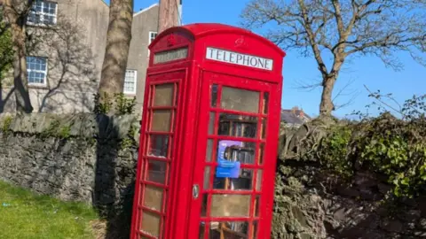 Denise Feenan A red phone box. A stone wall and trees are behind it and a large building can be seen in the background. Book are inside the phone box