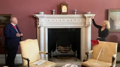 The Lord Mayor of York and Hannah Bellerby in the renovated drawing room at Mansion House. They stand either side of an ornamental white fireplace, behind two white chairs, and against a dark red painted wall.