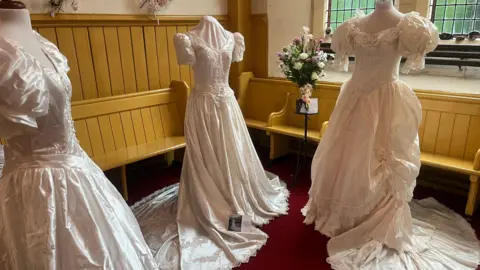 Three dresses on mannequins with church wooden seats behind them