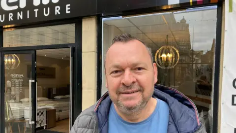 A man wearing a blue T-shirt and grey quilted jacket standing outside a shop front.