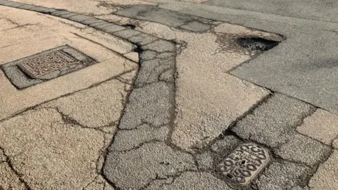 James Grant/BBC A road cover in large cracks virtually all over the tarmac. There are several potholes of varying depths and sections of tarmac in varying shades of black that have been recently patched up.
