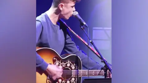 Side view of Tom A Smith playing a guitar, seen from waist height. He has short hair and is wearing a purple fleece. 
