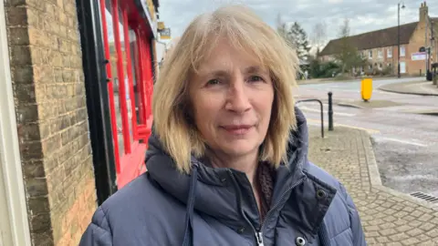 JOHN DEVINE/BBC Janet Taylor looking at the camera and slightly smiling. She is wearing a navy puffer jacket. Behind her is a street and buildings in Chatteris. 