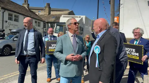 BBC/Aisha Iqbal Nigel Farage stands in Baildon, Bradford wearing a blue jacket. He's surrounded by Reform campaigners holding placards. He is talking to a man in a dark jacket wearing a Reform UK rosette.