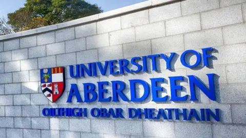 University of Aberdeen Bold blue lettering which says 'University of Aberdeen' on a grey brick wall.