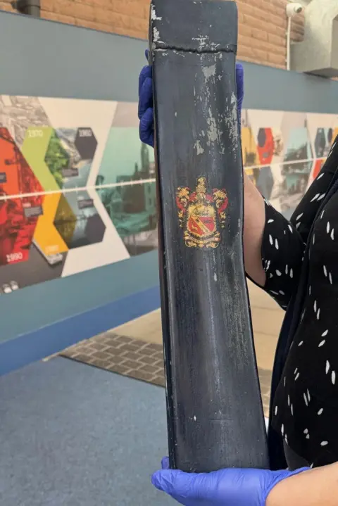 Barrow Police A wider shot of the time capsule which is long and narrow. It's being held up by a person who is wearing blue rubber gloves.