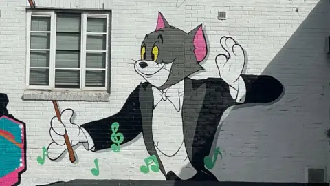 A street art mural featuring cartoon cat Tom, from Tom and Jerry, conducting music with green note symbols floating around,