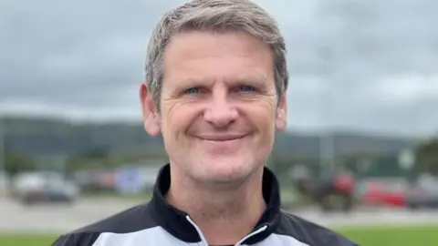 Nigel Osmond is smiling with grey hair. He is wearing a grey and black jacket with a zip.