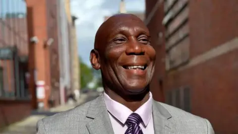 Sheffield Hallam University A man is smiling broadly, wearing a grey suit with a white shirt and purple striped tie