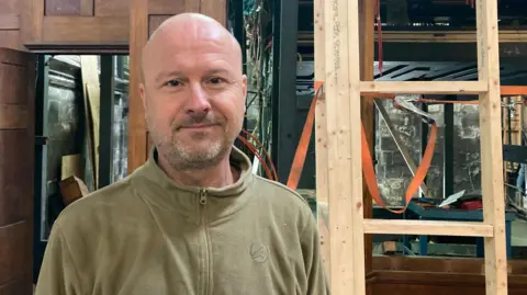 Marius Skjolaas - a man with a shaved head and stubble - stands in front of a jumble of metal and wooden structures. He is wearing a khaki-coloured fleece.