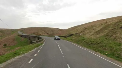 Google A road with grass verges either side and a car coming in the opposite direction. The road goes round a small corner.
