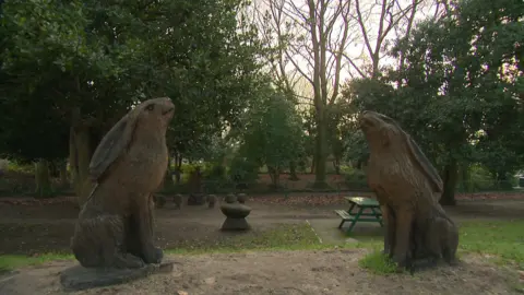 Two large wooden hare sculptures face each other, next to a path running through the park.