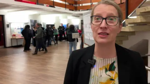 Sherilee Harper is looking at the camera, has blonde hair and blue eyes. She is wearing a cream blouse with a yellow and green flower on it, a black blazer and black framed glasses.