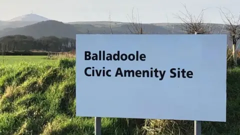 NORTHERN CiVIC AMMENITY SITE A white sign in a field with hills in the background on a sunny day. The sign reads Balladool Civic Ammenity Site.