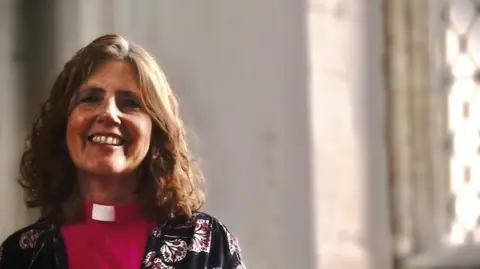 Brian Farmer/BBC Vicar Ruth Clay wear pink cassock and floral patterned cardigan 