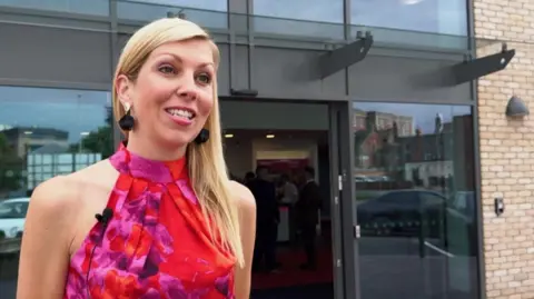 A woman with blond hair and wearing a smart red evening dress stands outside a new cinema building with an exterior dominated by large tinted windows.