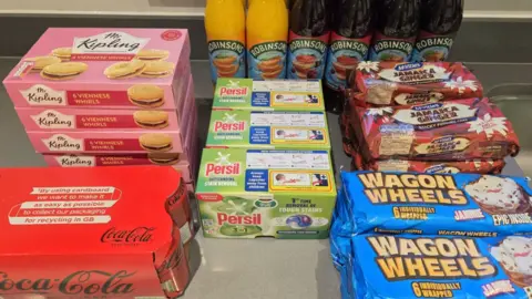 A multipack of Coca-Cola, six packs of Mr Kipling Viennese Whirls, six bottles of Robinsons squash, four packs of McVities Jamaica Ginger cakes, several packs of Wagon Wheel and three boxes of Persil washing tabs are arranged in a display of Peter's discounted Co-op Deliveroo order.