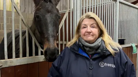 Grace Muir has long blonde hair. She is stood next to a dark coloured horse in a stable with metal bars.