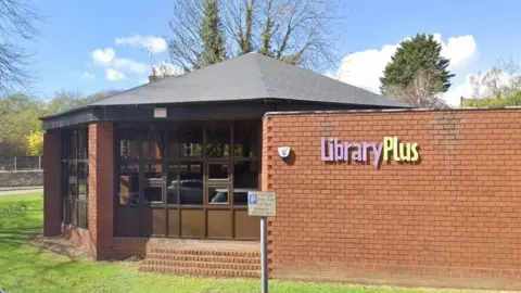 Brick-built hexagonal single-storey library building with many windows. A brick to the right has the words Library Plus in pink and yellow lettering. There are trees and grass around the library.