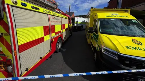 Blue-and-white police tape stretches across a road, behind which is a bright red fire engine, a yellow ambulance, and behind both of these, a white bomb squad van with its rear doors open.