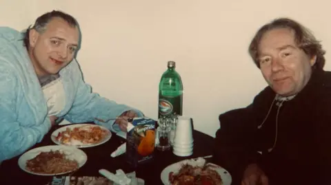 Bob Baldwin Rik Mayall, his hair slicked back, sits at a table eating what looks like a curry. He is wearing a fluffy baby blue dressing gown. On the other side of the table is a middle-aged man with floppy hair with a plate of food in front of him. Cups, a bottle and a carton of orange juice are also on the table.