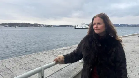 A woman in a brown coat stands looking over water on a cold, grey day in Oslo