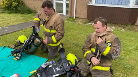 IOM Fire and Rescue Service Two firemen remove their equipment in a garden. 