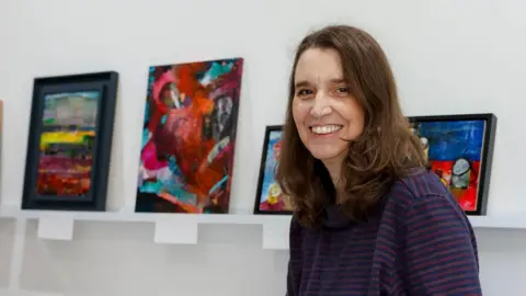 Ellie McCallum Photography A local artist smiling at the camera displaying her artwork at the Royal United Hospitals (RUH) in Bath. 