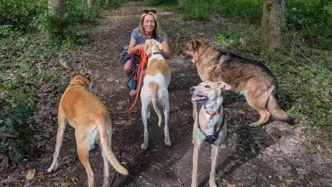 A woman with blonde hair crouches on a woodland path as she strokes a beige and white dog on a lead. She is also holding the lead to a brown lurcher. A greyhound in a harness is facing the camera. A German Shepherd is looking towards the woman, who is wearing a grey v-neck t-shirt and shorts, has sunglasses pushed back on her head, and is smiling.