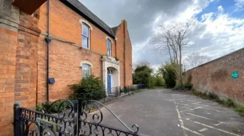 Somerset Council Image shows the side of a red brick house with a blue door and a large tarmac area in front of it. 