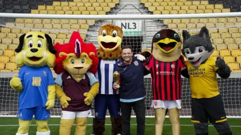 Mark Rhodes stands in a goal at the Molineux stadium with five football mascots: a dog, a lion, two birds, and a wolf. 
