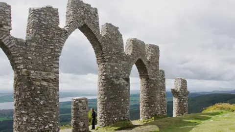 Julian Paren/Geograph A person stands under an arch of the monument built to resemble the ruined gates of Negatapam, a town in India. The person is taking in the view over the Cromarty Firth. 