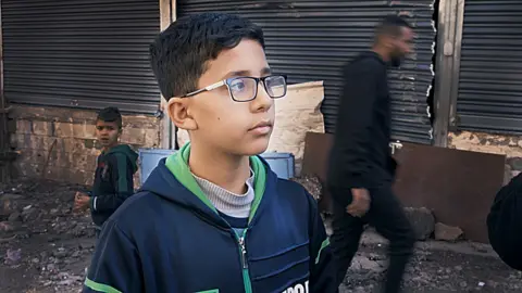 Haytham, a 12-year-old Palestinian-Canadian, wearing glasses and a zip-up hoodie, standing on the street with others walking by