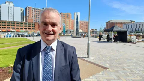 Chris Conway is smiling. He's wearing a navy suit with a white shirt and a blue patterned tie. A large sculpture is in the background. 
