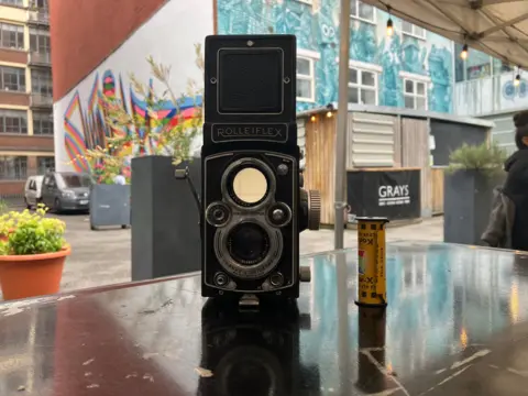 A Rolleiflex camera and a reel of film sit on a table. There is a colourful mural in the background.