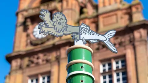 The lion head and herring tail emblem from the Great Yarmouth Borough Council heraldic crest, adorns the top of a standard outside the Great Yarmouth Town Hall, which is featured in soft focus. The building is made of red brick, and red masonry, with white windows, decorative features and a clock, plus white-painted window frames. The standard upon which the sign is mounted, is green.