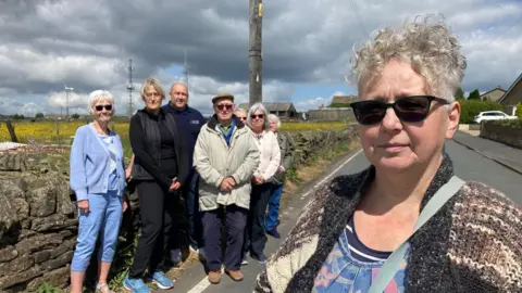 A woman with grey, curly hair on the right and seven men and women in a group behind her all standing on a road.