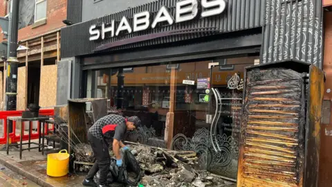 A restaurant called SHABABS is charred from a fire, with a person clearing up the mess at the front.