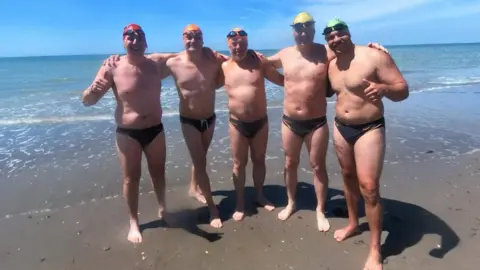 Five men wearing small black swimming pants, caps and goggles have their arms round each other and are smiling for the camera. They are standing on a beach in front of the sea.