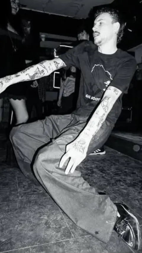 Ben Keen A black and white image of a young man dancing. He is crouched low, with his ankles out-turned. He wears formal trousers, black loafers and a black graphic T-shirt. He has two sleeves of tattoos on his arms. Behind him is a dancefloor filled with white powder.