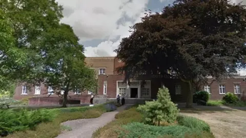 Google An old brick building with white windows, with trimmed hedges and trees outside