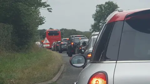 BBC A long traffic jam of cars and buses. There is a road sign on the grass verge. 