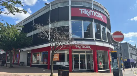 The store has TK Maxx in white above the front door and also higher up in the same area of the building on the outside. A no-entry traffic sign is visible in front of the building.