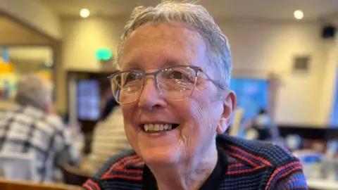 Elaine Hodgson smiling at someone past the camera, with a blurred background of a pub or social club. She has very short grey hair. She is wearing glasses and a black and blue striped top, with some red stripes.