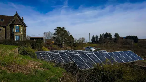 A row of solar panels surrounded by a hillside garden with a stone house at the top