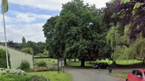Google Part of a public park, featuring lots of fully leafed trees, a footpath, grassed areas and a noticeboard. A red car is parked, partially in shot to the right of the picture.