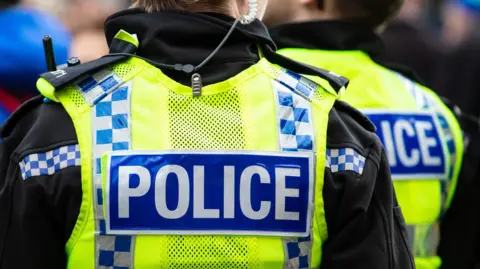 Two police officers wearing yellow hi-vis vests with "police" written on the back. One officer has an earpiece. They both have their backs to the camera.