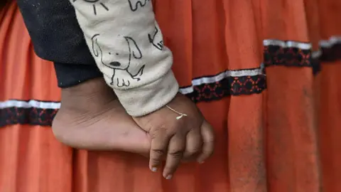 A close-up of a small child’s bare foot and hand is shown against a backdrop of their mother's skirt - a traditional indigenous outfit. It appears the child is being held by their mother. The child is wearing a light-coloured sleeve printed with a simple cartoon dog, and the hand hangs loosely beside the foot.
