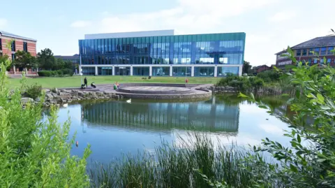 A view between plants of the Central Square pond. The weather is bright and lighting up a glass building on the other side of the pond. 